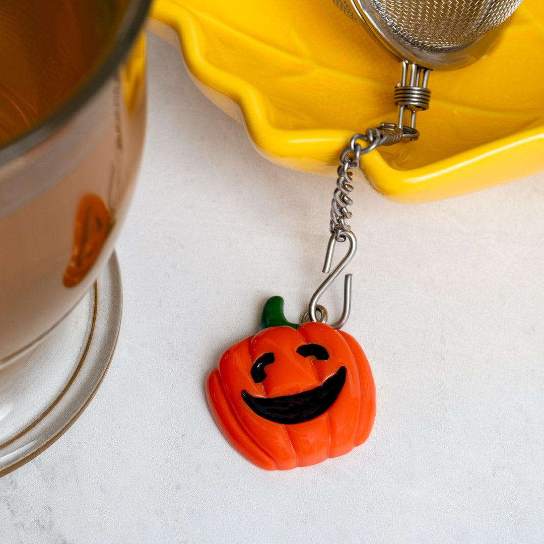 Pumpkin charm hanging from tea ball infuser next to steeped tea and on yellow tea rest.